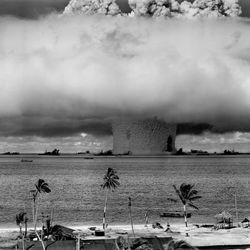 A black and white photo showing the enormous mushroom cloud caused by the nuclear explosion. The photo was taken from a distant island shoreline, the sandy beach of which is visible in the foreground. The mushroom cloud is set on the horizon and takes up most of the frame. 