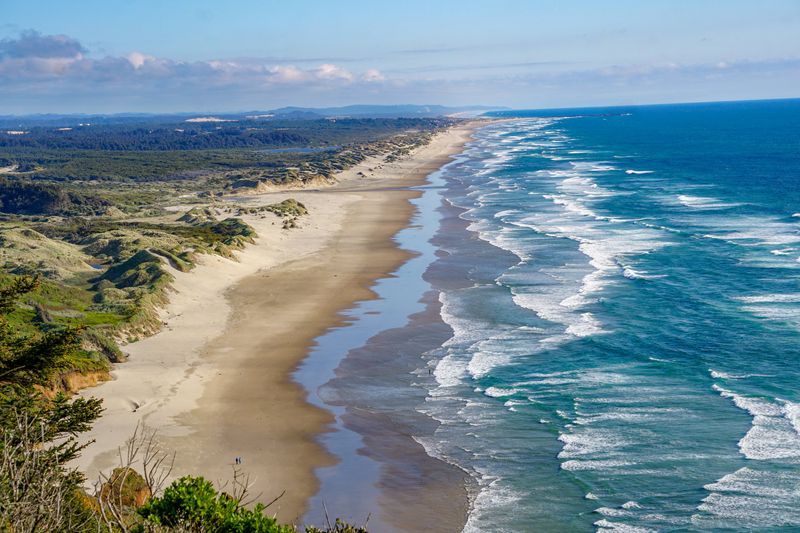 A photo showing the coast of Oregon from an elevated point. A sandy beach is visible on the left of the shot and extends all they way to the horizon point. There are small break water waves coming in from the right. 
