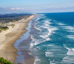 A photo showing the coast of Oregon from an elevated point. A sandy beach is visible on the left of the shot and extends all they way to the horizon point. There are small break water waves coming in from the right. 