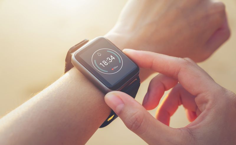 A person adjusts a black smartwatch on their wrist, displaying the time 18:34 on its screen.