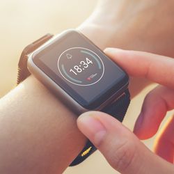 A person adjusts a black smartwatch on their wrist, displaying the time 18:34 on its screen.