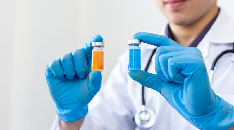 close up of doctor wearing blue gloves holding two vials of liquid, one yellow and one blue, one in each hand