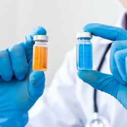 close up of doctor wearing blue gloves holding two vials of liquid, one yellow and one blue, one in each hand